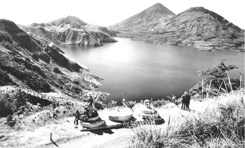 Fotografía antigua en blanco y negro del lago de Atitlán: el agua rodeada de volcanes y laderas, vista desde lo alto de un camino de tierra. En primer plano, varias personas con sombrero junto a unas grandes piedras de molino.